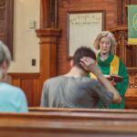 An elderly female clergyperson leads a service inside a church, engaging with the congregation.